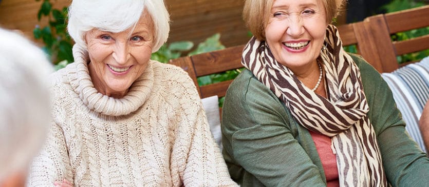Senior women with charming smiles listening to their male friend with interest while having gathering at cozy small patio