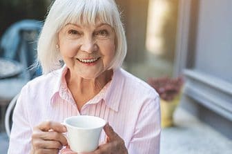 Waist up portrait of happy elderly woman sitting at table in cafe and holding cup of tea. She is enjoying evening on her own. Copy space in right side